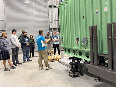 FRIB Outreach Coordinator Zach Constan speaks to a group of students in front of a green cryomodule in a truck bay at FRIB.