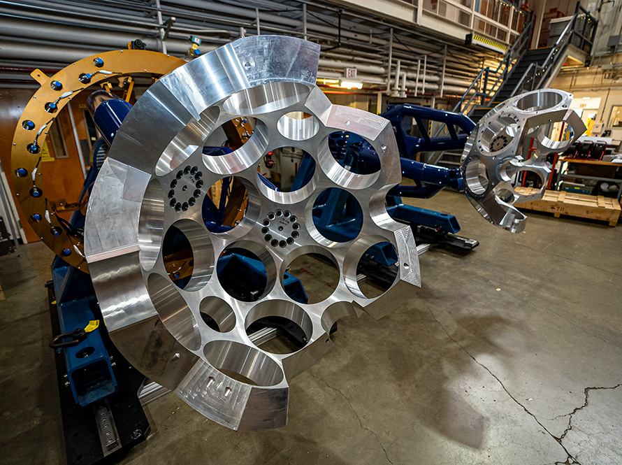 Two large metallic structures with multiple round machined holes in a radial pattern. The structure is mounted on a blue support frame inside an industrial lab space.