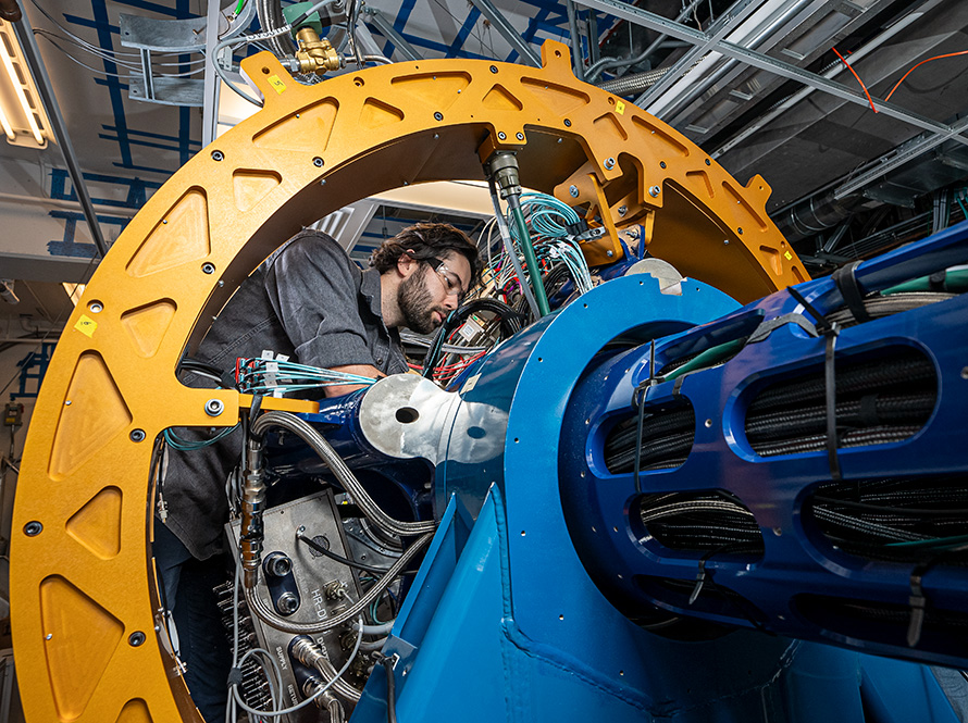 A person looks at something hidden behind GRETA’s cylindrical blue metal structure, which holds numerous cables. The person is framed by a large, circular yellow arch that connects to the blue assembly. 