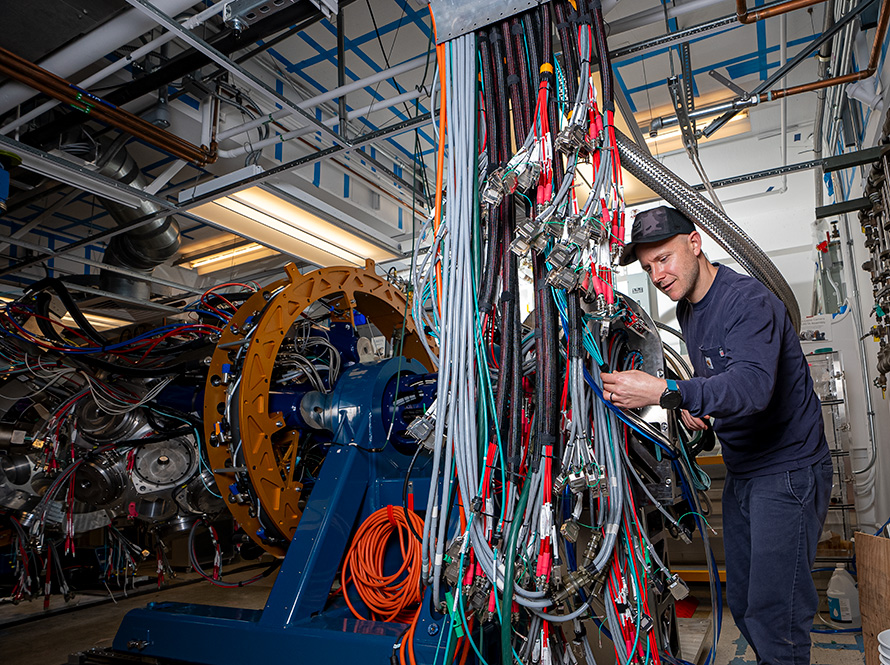 A person holds one of dozens of cables neatly draped from a tray several feet in the air. GRETA’s frame, fully instrumented with detectors and cables, is visible in the background.