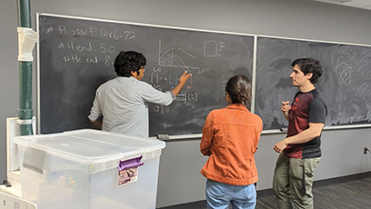 Three students have a discussion while facing a chalkboard. In the foreground on the left is the “marble nuclei fragmentation box”—a demonstration device used to explain how rare isotopes are produced at FRIB in a simplified way.  