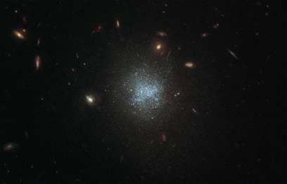 A dark telescope image is dotted with small, yellow spiral and disc-shaped galaxies. At its center, though, the galaxy shown looks like a dispersed cloud of blue dots.
