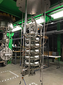 Niobium superconducting elliptical cavity (long piece of silver scientific equipment suspended from the ceiling) for FRIB Upgrade to 400 MeV/u capable to provide 12.4 MV accelerating voltage.