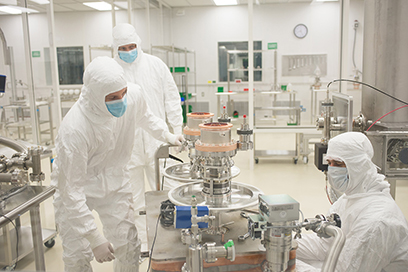 Three graduate students wearing masks and white protective suits work on scientific instruments in the cleanroom.