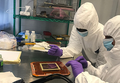 Two graduate students wearing white protective suits, purple gloves, and masks work together at a table in the laboratory.