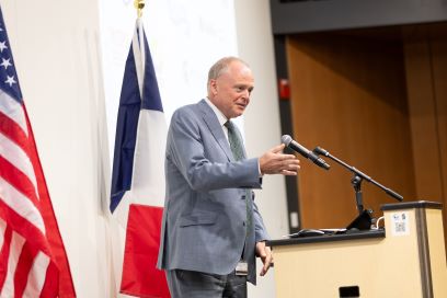 Facility for Rare Isotope Beams Laboratory Director Thomas Glasmacher speaks into a microphone at a podium. An American flag is in the foreground (left) and a French flag is behind him.
